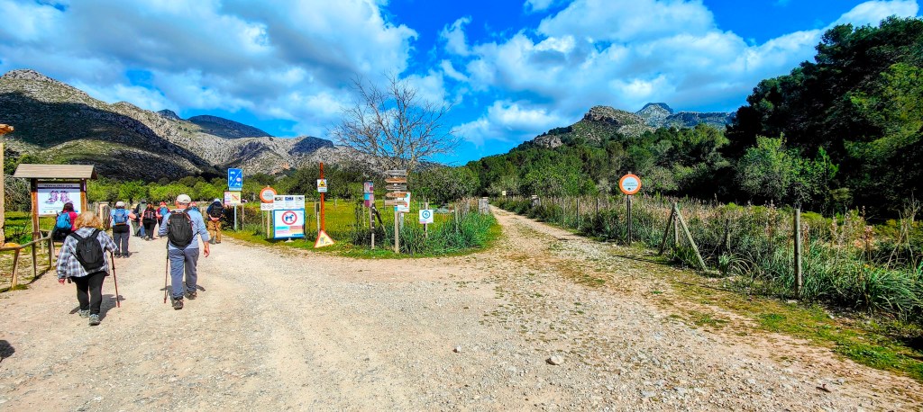 Grupo de personas caminando por un sendero en una zona montañosa, con señales de información y advertencia a lo largo del camino.