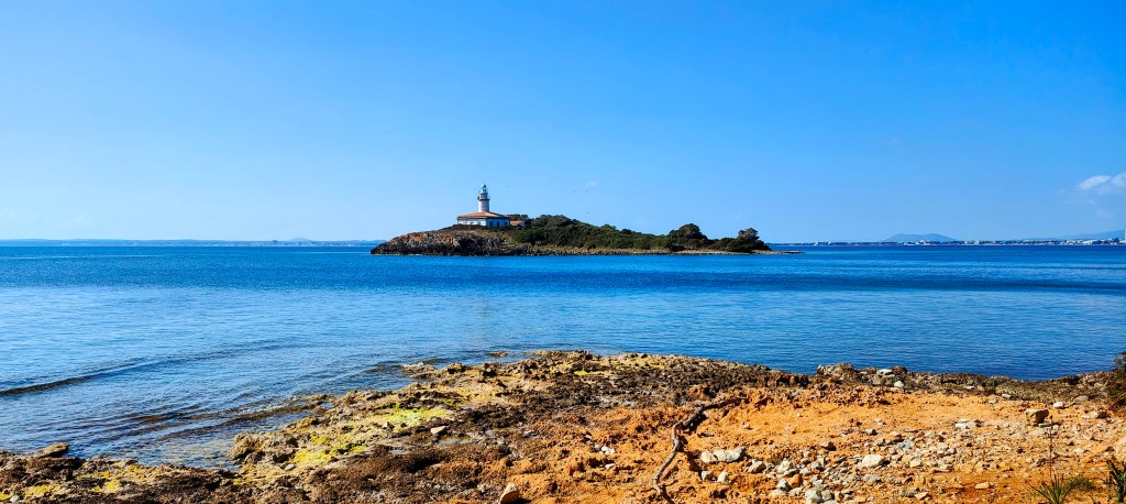 Vista de un faro en una isla, rodeado de agua azul y un cielo despejado.