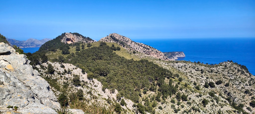 Vista panorámica de montañas y costa, con árboles verdes y mar en el fondo, bajo un cielo despejado.