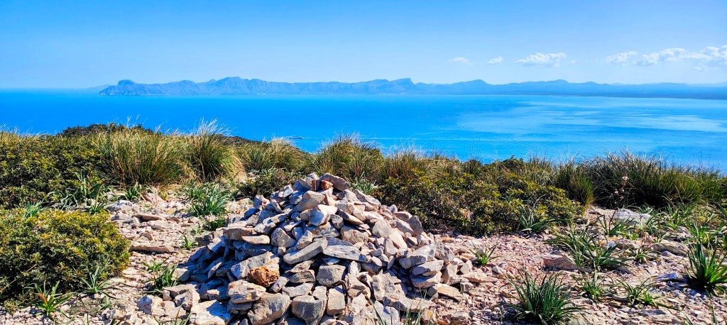 Montículo de piedras en un paisaje costero con mar azul y montañas al fondo.