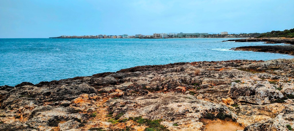 Vista de la costa con rocas en primer plano y un fondo de ciudad sobre el mar.