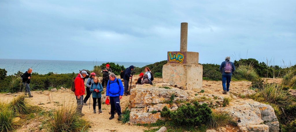 Un grupo de personas en una colina cerca del mar, con un monumento de concreto marcado con graffiti que dice 'Love'.