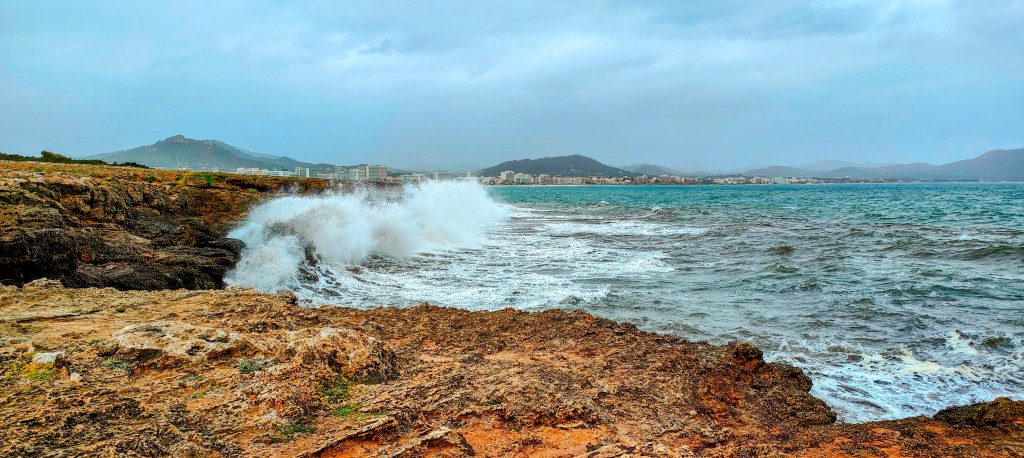 Vista de un paisaje costero con olas rompiendo sobre las rocas, cielo nublado y montañas al fondo.
